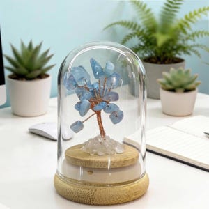Op de afbeelding: Een decoratieve boomsculptuur met lichtblauwe edelstenen als bladeren, koperdraad takken en een houten stam, ingekapseld in een glazen koepel. De basis is gemaakt van hout en staat op een wit oppervlak. De achtergrond bevat potplanten.