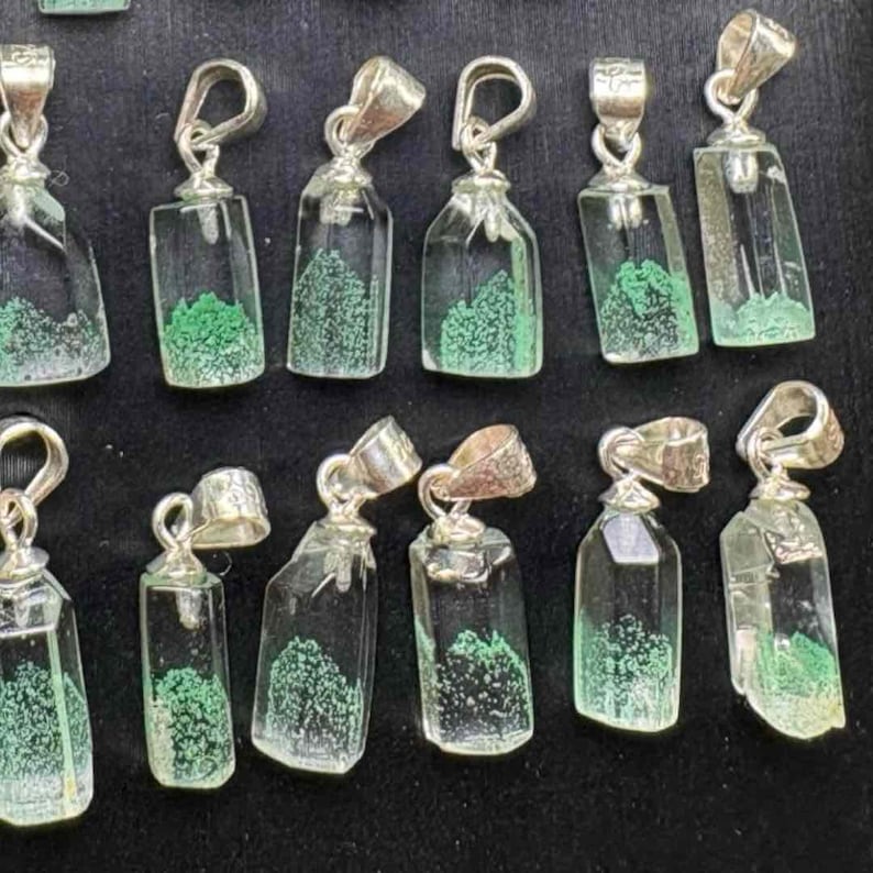 May include: A collection of clear glass pendant necklaces with silver-colored metal loops. Each pendant contains a green, textured, tree-like design. The pendants are displayed on a black surface.