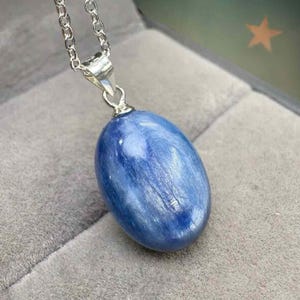 Può includere: Una collana con pendente ovale blu e catena d'argento. Il pendente è una pietra liscia e lucida con aspetto marmorizzato. La collana è esposta su una superficie grigia.