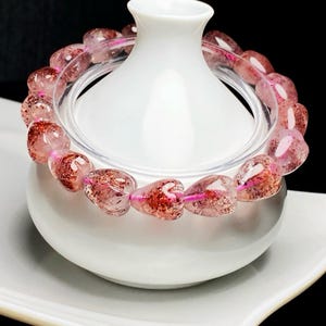 May include: A clear quartz bracelet with red inclusions and a pink string. The beads are oval-shaped and strung together. The bracelet is displayed on a white ceramic container.