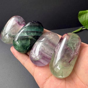 May include: Four polished, oval-shaped fluorite stones featuring translucent bands of purple, green, and clear mineral inclusions. Each stone has a smooth, glossy surface that reflects light, showcasing natural colour gradients and internal crystalline patterns.