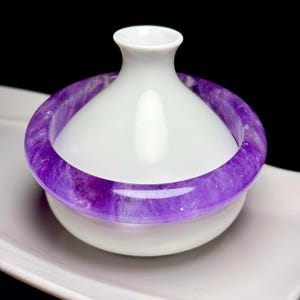 May include: A smooth white ceramic vessel with a tapered neck and a wide base, featuring a translucent purple amethyst bangle bracelet encircling the midsection of the container. The bracelet displays natural crystalline textures and deep violet hues.