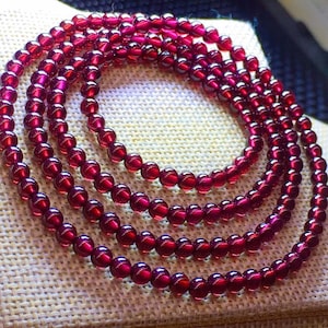 May include: Multiple strands of a deep red beaded necklace. The beads are round and have a glossy finish, reflecting light. The necklace is arranged in a circular pattern, showcasing the rich color and texture of the gemstones.