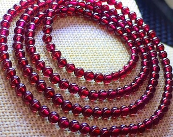 May include: Multiple strands of a deep red beaded necklace. The beads are round and have a glossy finish, reflecting light. The necklace is arranged in a circular pattern, showcasing the rich color and texture of the gemstones.