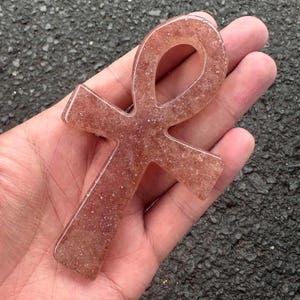 May include: An ankh symbol crafted from a translucent, reddish-pink material with a glittery, crystalline texture throughout. The object features a smooth, polished surface and a distinct cross shape with a looped top section, held in a palm.