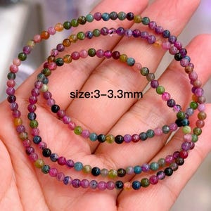 May include: Three strands of colorful gemstone bead necklaces. The beads are small, ranging from 3 to 3.3 mm in size, and display a variety of colors including pink, green, blue, and orange. The necklaces are displayed on a light-toned background.
