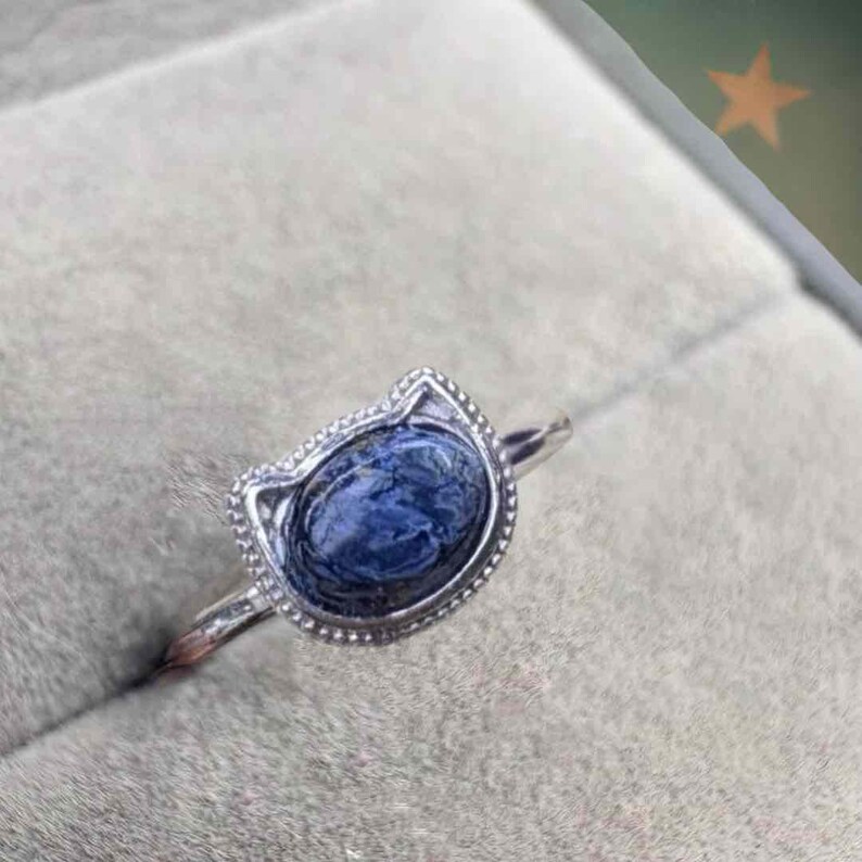 May include: A silver ring featuring a blue oval gemstone set in a cat-shaped bezel. The ring has a delicate band and a textured border around the gemstone. The gemstone has a marbled appearance.