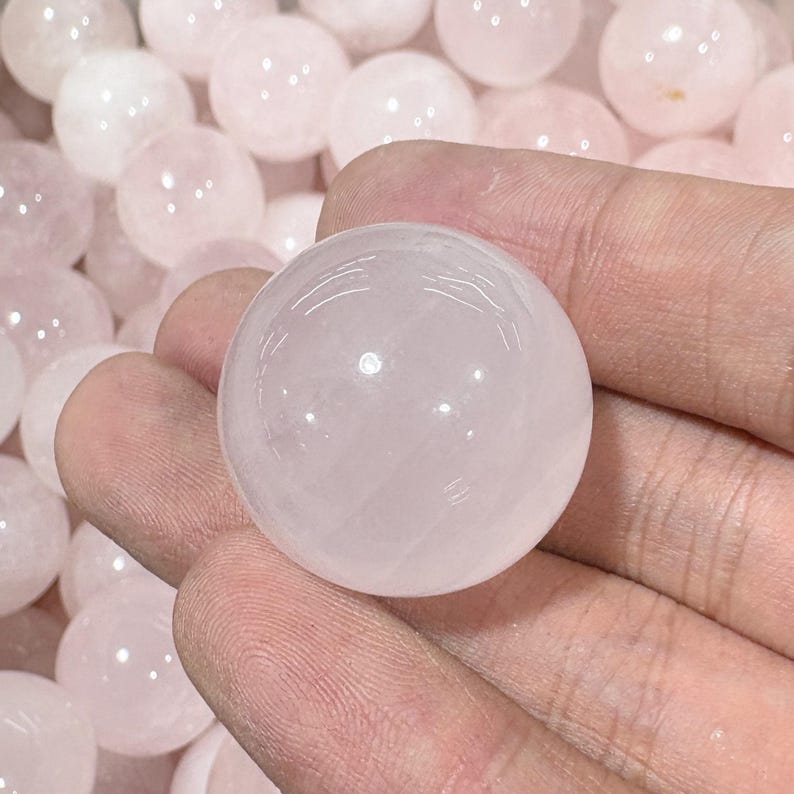 May include: A collection of polished rose quartz spheres. One sphere is held in a hand, showcasing its translucent pink color and smooth surface. The background is filled with similar spheres, creating a soft, diffused effect.