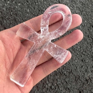 May include: A clear, translucent quartz crystal carved into the shape of an ankh symbol. The object features a smooth, polished surface with visible internal fractures and natural inclusions throughout the transparent, colourless stone structure.