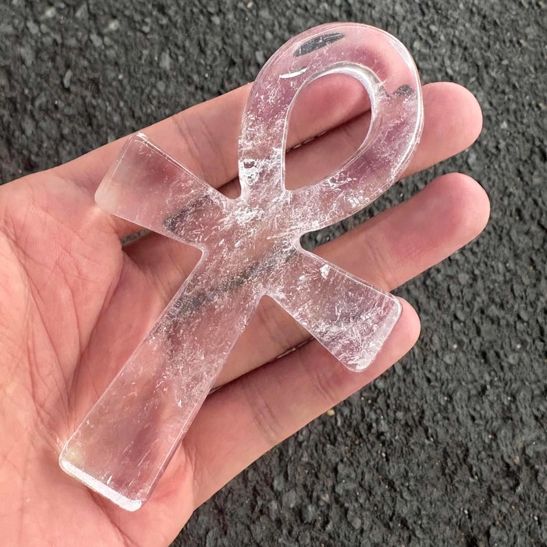 May include: A clear, translucent quartz crystal carved into the shape of an ankh symbol. The object features a smooth, polished surface with visible internal fractures and natural inclusions throughout the transparent, colourless stone structure.