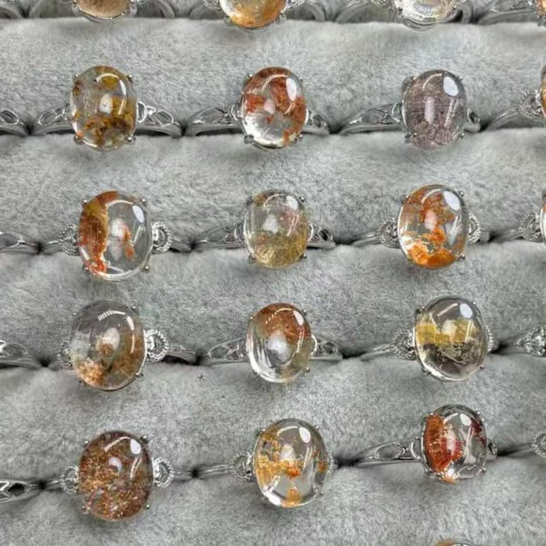 May include: A selection of silver rings, each featuring an oval, clear gemstone with reddish-brown inclusions. The rings are presented on a grey, textured surface, highlighting the diverse designs and colours.