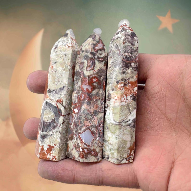 May include: Three polished stone obelisks with a mix of colors including red, cream, and gray. Each obelisk has a faceted shape and a small, clear cap at the top. The stones are held in a hand.