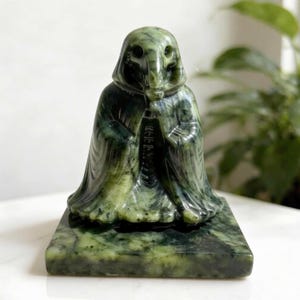 Op de afbeelding: Een uit groene steen gesneden beeldje van een figuur met een kap en een lange, slurfachtige neus, staand op een vierkante, gepolijste groene stenen voet. Het beeldhouwwerk vertoont donker- en lichtgroene marmerpatronen over het gladde oppervlak.