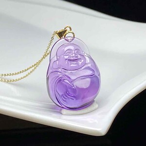 May include: A purple pendant necklace featuring a smiling Buddha figure. The pendant is made of a translucent, polished material and is suspended from a delicate gold chain. The Buddha figure is carved with detailed features.