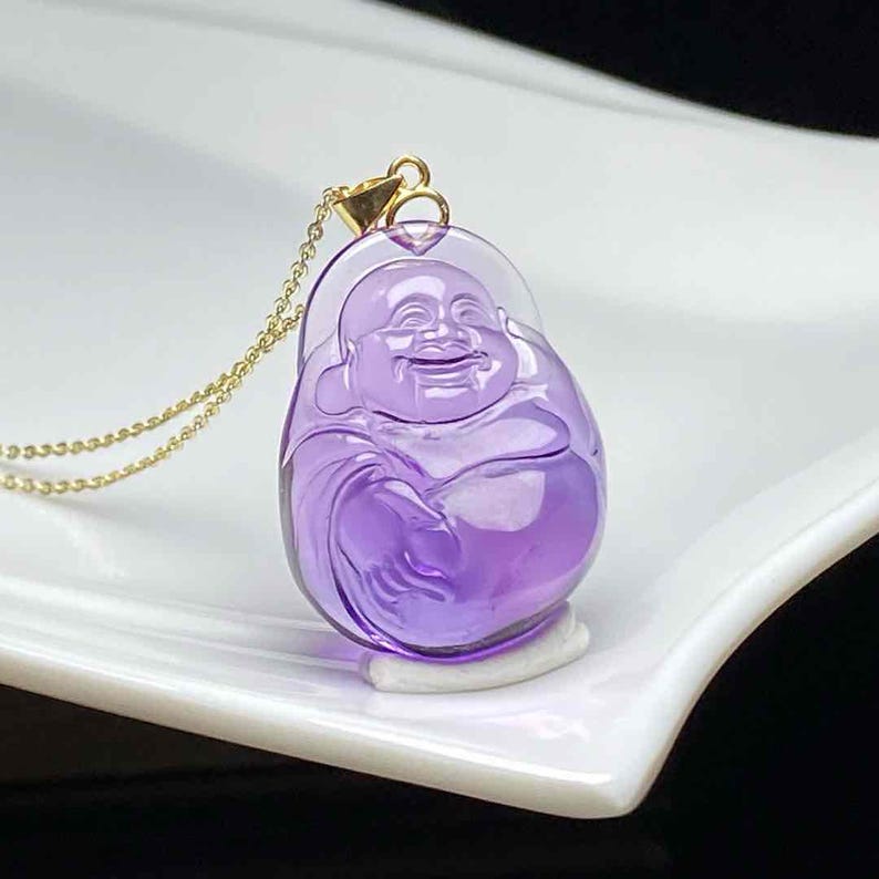 May include: A purple pendant necklace featuring a smiling Buddha figure. The pendant is made of a translucent, polished material and is suspended from a delicate gold chain. The Buddha figure is carved with detailed features.