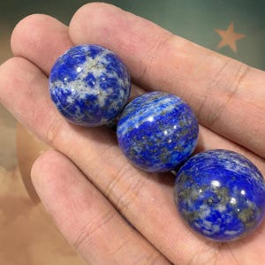 May include: Three polished lapis lazuli spheres, each approximately 3.8 cm in diameter, are displayed. The spheres exhibit a deep blue hue with white and gold flecks, showcasing the natural patterns of the stone. The smooth, round gemstones are held in a hand.