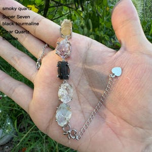 May include: A silver bracelet featuring a series of natural gemstones, including smoky quartz, Super Seven, black tourmaline, clear quartz, and fluorite. The bracelet has a delicate chain and a heart-shaped clasp. The gemstones are in various colors.