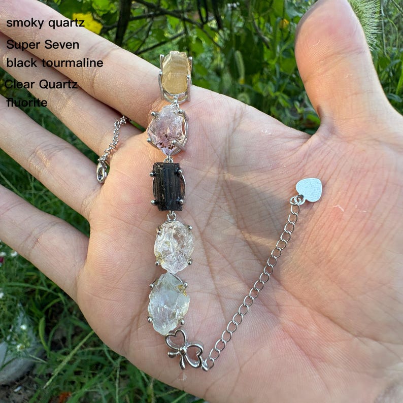 May include: A silver bracelet featuring a series of natural gemstones, including smoky quartz, Super Seven, black tourmaline, clear quartz, and fluorite. The bracelet has a delicate chain and a heart-shaped clasp. The gemstones are in various colors.