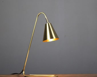 May include: A gold-colored desk lamp with a conical shade and a curved arm. The lamp is positioned on a wooden surface, with a dark gray background. The lamp is plugged into an electrical outlet.