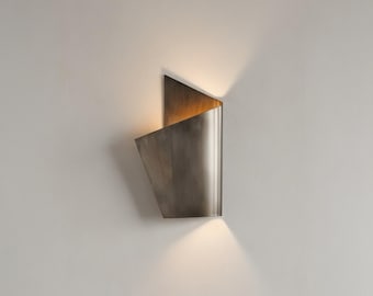 May include: A modern wall sconce with a metallic finish. The fixture has a geometric design with a triangular top and a curved, folded body. The sconce emits a warm light, casting a soft glow on the wall below.