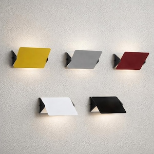 May include: Five modern wall sconces in yellow, grey, red, white, and black. Each sconce has a rectangular shade with a folded design, mounted on a black bracket. The sconces are mounted on a textured, off-white wall.