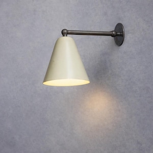 May include: Cream-colored wall sconce with a conical shade and a dark metal arm and mounting plate. The light is on, casting a warm glow on the wall. The sconce is a modern design.