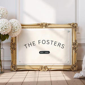 May include: A framed canvas with the text "THE FOSTERS" and "EST. 1981" in black. The frame is ornate gold, and the canvas is cream-colored. A vase of white flowers sits to the left, and the background is a white wall and wooden floor.