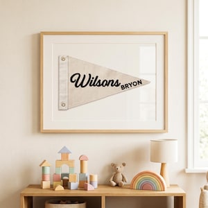 Op de afbeelding: Een ingelijste wimpel met de naam "Wilsons" in een sierlijk lettertype en de naam "BRYON" in een kleiner lettertype. De wimpel is lichtbeige en is gemonteerd op een witte achtergrond. De lijst is van lichtkleurig hout. De afbeelding staat boven een houten plank met speelgoed.