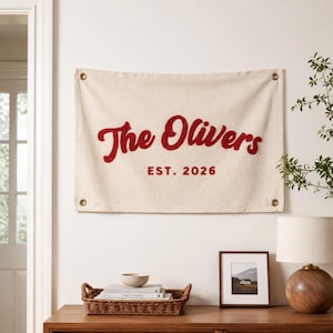 May include: A rectangular off-white canvas wall banner featuring the red embroidered text The Olivers and EST. 2026. The banner has four brass-colored metal grommets, one in each corner, for hanging purposes. The fabric has a textured, woven appearance.