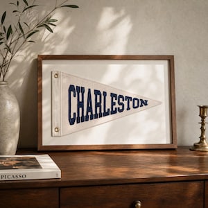 Op de afbeelding: Een ingelijste wimpel met het woord "CHARLESTON" in marineblauwe letters. De wimpel is wit en heeft twee messing ogen. De lijst is van donker hout. De wimpel wordt op een houten oppervlak getoond.