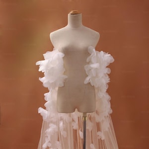 Puede incluir: Una estola de novia blanca, presentada sobre un maniquí. Está adornada con grandes acentos blancos y esponjosos en forma de plumas. La falda es transparente y fluida. El fondo es de color neutro y cálido.