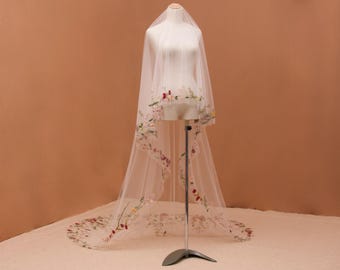 May include: A long, sheer white bridal veil draped over a mannequin, featuring delicate, multi-colored floral embroidery along the scalloped edges. The embroidery includes small red, pink, and green botanical motifs that extend along the hemline.