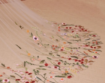 May include: A sheer, off-white tulle veil featuring delicate, multi-colored floral embroidery. The design showcases various wildflowers in shades of red, pink, yellow, and green, scattered across the fabric in a trailing, organic pattern.