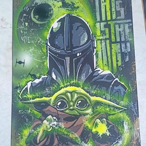 Puede incluir: Una obra de arte enmarcada que presenta a The Mandalorian y Grogu de Star Wars. La obra tiene un fondo oscuro con acentos verdes y amarillos. El texto "THIS IS THE WAY" es visible. El casco del Mandaloriano y la cara de Grogu son prominentes.