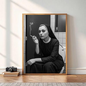 May include: Black and white framed print of a person in a nun's habit, smoking a cigarette while seated on a toilet. The image is framed in a light brown wooden frame. A camera and books are in the foreground.
