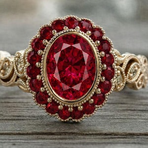 Pode incluir: Um anel dourado com uma pedra preciosa vermelha oval e facetada no centro, rodeada por pedras vermelhas menores. A banda apresenta detalhes intrincados de filigrana e volutas, criando um design ornamentado e elegante.