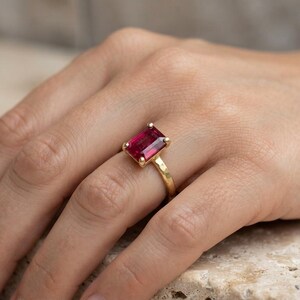 May include: A gold ring featuring a rectangular, emerald-cut deep pink gemstone set in a four-prong setting. The band of the ring has a textured, hammered finish, providing a distinct surface appearance compared to a smooth, polished metal band.