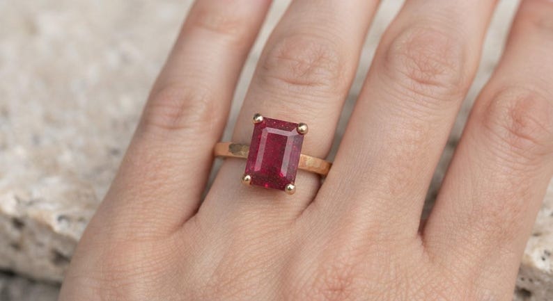 May include: A gold ring featuring a rectangular, emerald-cut deep red gemstone set in a four-prong setting. The band of the ring has a textured, hammered finish, providing a distinct surface appearance compared to a smooth, polished metal band.