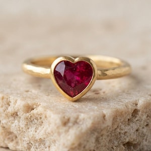 Puede incluir: Un anillo de oro con una piedra preciosa de color rojo intenso en forma de corazón engastada en un bisel. La banda del anillo tiene un acabado texturizado y martillado, creando un contraste con la superficie lisa y pulida de la piedra.