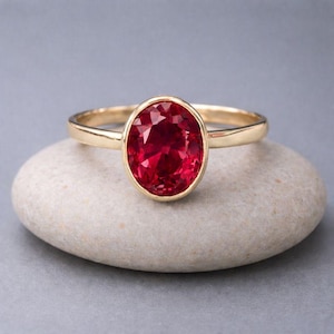 May include: A gold ring featuring a large, oval-cut, deep red gemstone. The gemstone is set in a bezel setting, and the band is a simple, polished gold. The ring is displayed on a smooth, light-colored stone, against a soft gray background.
