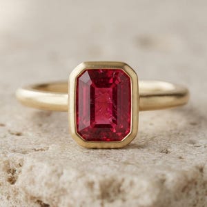 May include: A gold ring featuring a rectangular, emerald-cut, deep red gemstone set in a smooth bezel setting. The band is a simple, polished gold metal. The gemstone is centered and displays a vibrant, saturated red hue with clear facets.
