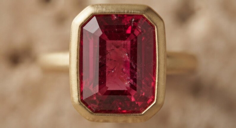 May include: A ring featuring a large, rectangular, faceted deep pink gemstone set in a smooth, brushed yellow gold bezel setting. The stone displays internal inclusions and a vibrant, saturated colour, while the gold band has a matte finish.