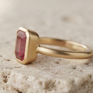May include: A gold ring featuring a rectangular, emerald-cut red gemstone set in a smooth, bezel-style mounting. The band has a matte, brushed finish, and the gemstone is positioned vertically, showcasing its deep red colour and faceted surface.