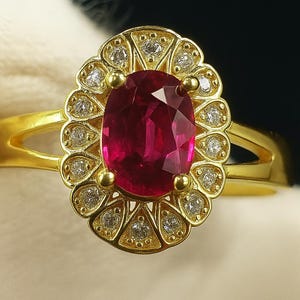 May include: A gold ring featuring a large, oval-cut ruby gemstone. The ruby is surrounded by a halo of smaller diamonds set in a gold filigree design. The ring's band is a simple, polished gold.