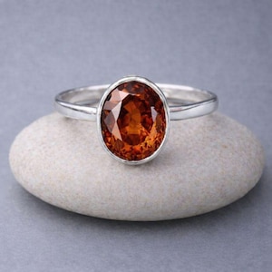 May include: A silver ring featuring a large, oval-cut, orange gemstone. The gemstone is set in a simple, polished silver band. The ring is displayed on a smooth, light-colored stone against a gray background. A piece of jewelry.