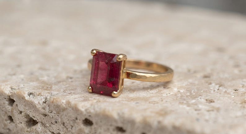 May include: A gold ring featuring a rectangular emerald-cut deep red gemstone set in a four-prong setting. The band of the ring has a textured, hammered finish, providing a distinct surface appearance compared to a smooth polished metal band.