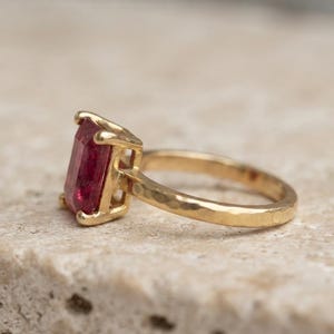 May include: A gold ring featuring a rectangular, emerald-cut red gemstone set in a four-prong setting. The ring band has a distinct hammered texture, providing a reflective surface that complements the deep red colour of the central stone.