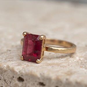 May include: A gold ring featuring a rectangular emerald-cut deep red gemstone set in a four-prong setting. The band of the ring has a textured, hammered finish, providing a distinct surface appearance compared to a smooth polished metal band.