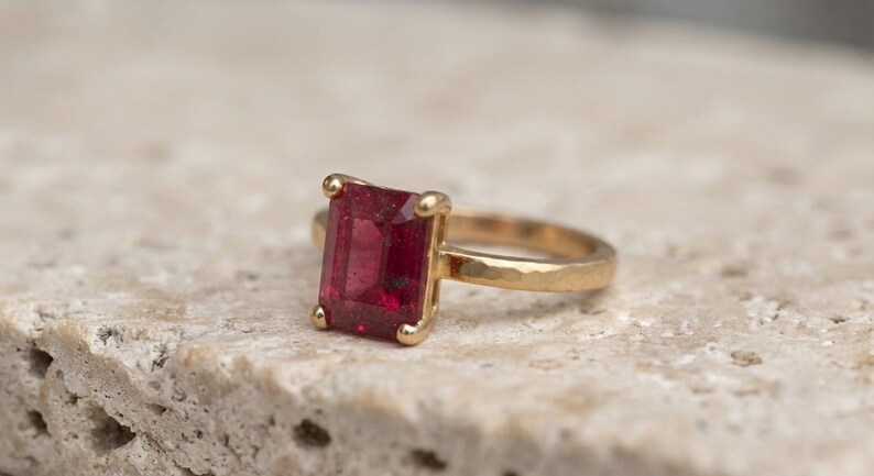 May include: A gold ring featuring a rectangular emerald-cut deep red gemstone set in a four-prong setting. The band of the ring has a textured, hammered finish, providing a distinct surface appearance compared to a smooth polished metal band.