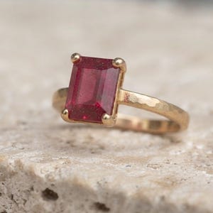 May include: A gold ring featuring a rectangular emerald-cut red gemstone set in a four-prong setting. The band of the ring has a distinct hammered texture, providing a matte finish to the metal surface that contrasts with the faceted red stone.